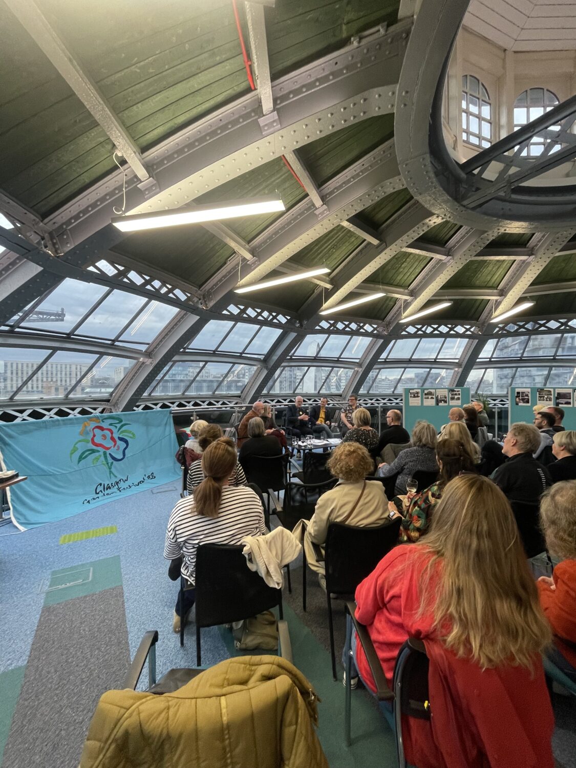 A group of people at a live podcast recording. There is a Garden Festival flag on display.